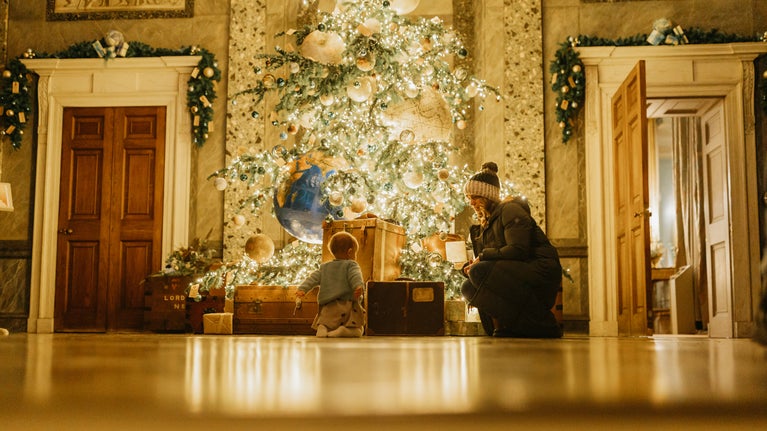 A warmly lit room with a large, decorated Christmas tree in the center, surrounded by vintage suitcases and boxes. A child in light-colored pajamas sits on the floor facing the tree, while an adult in a winter hat and coat crouches beside them. Wooden doors on either side are framed with festive garlands